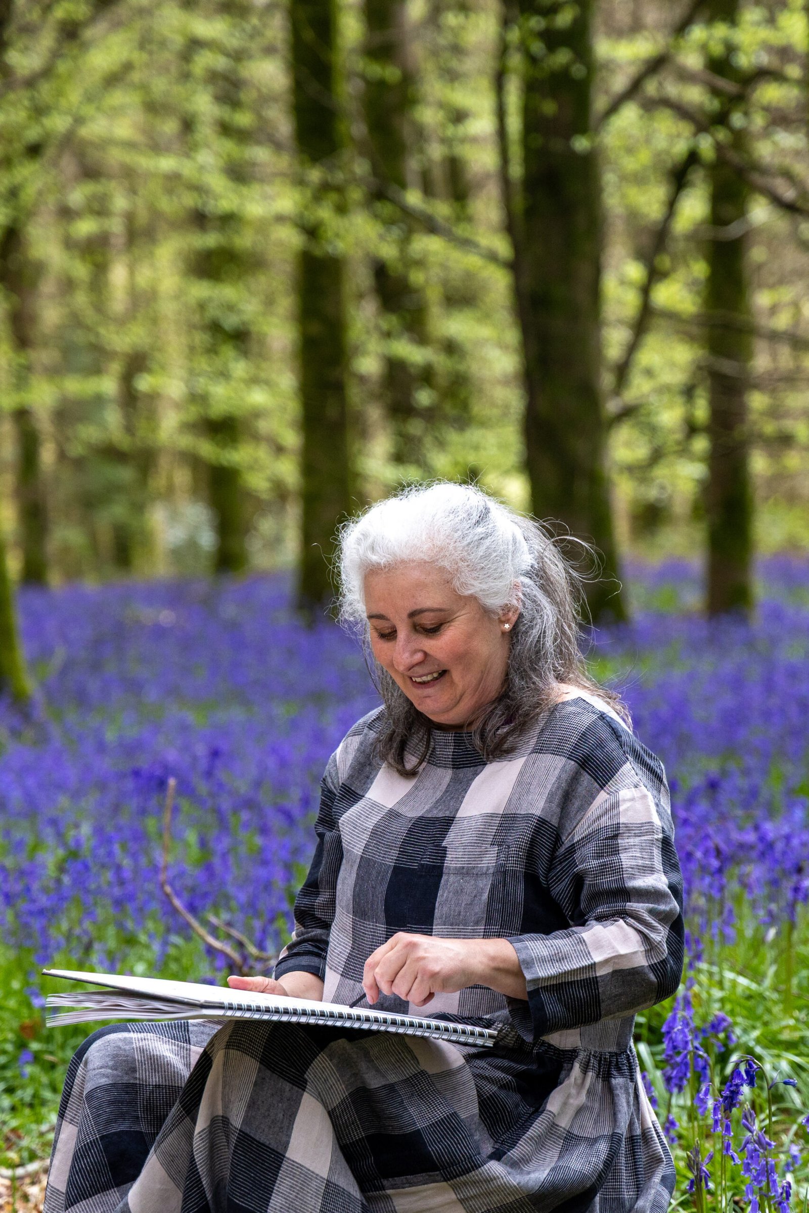 Arty Genny sitting amongst bluebells with a sketchbook on her lap, sketching with charcoal; a mindful art experience