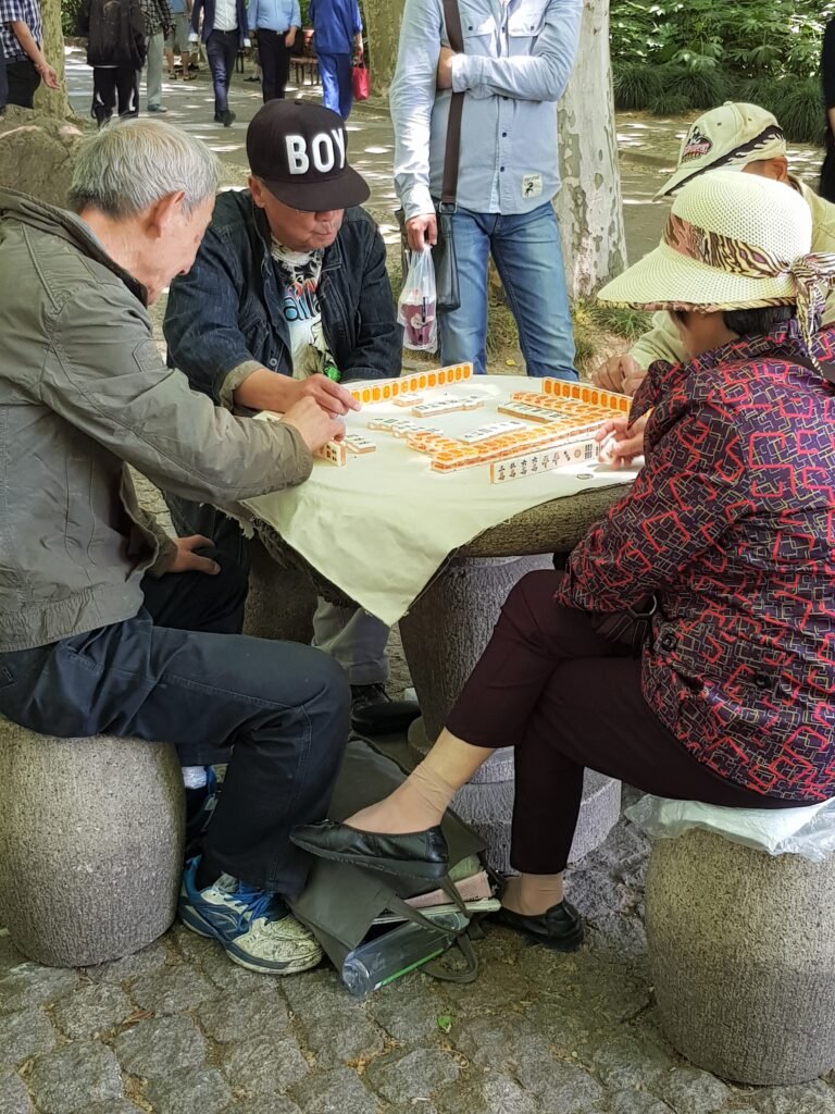 Four Chinese adults sitting round a stone table in Renmin Park shaded by trees; the table is covered with a white cloth and they are playing Mah Jong
