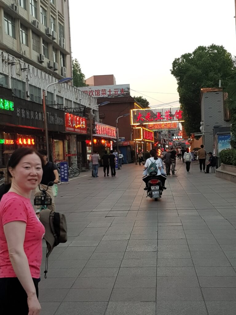 A view of a street in China at dusk. It's a pedestrian area and the street is called "Gourmet Street". There are bright lights on all the buildings and people walking up and down the street, as well as someone riding on a moped