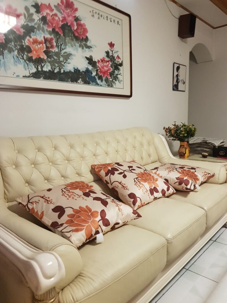 A cream leather in the foreground with three floral-design cushions loosely arranged. On the wall behind the sofa hangs a large Chinese painting of a bright pink peony. The walls are painted off-white