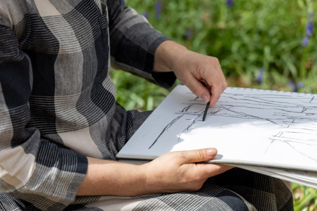 A close-up of Genny calmly sketching outdoors with charcoal into a sketchbook that is balanced on her lap.