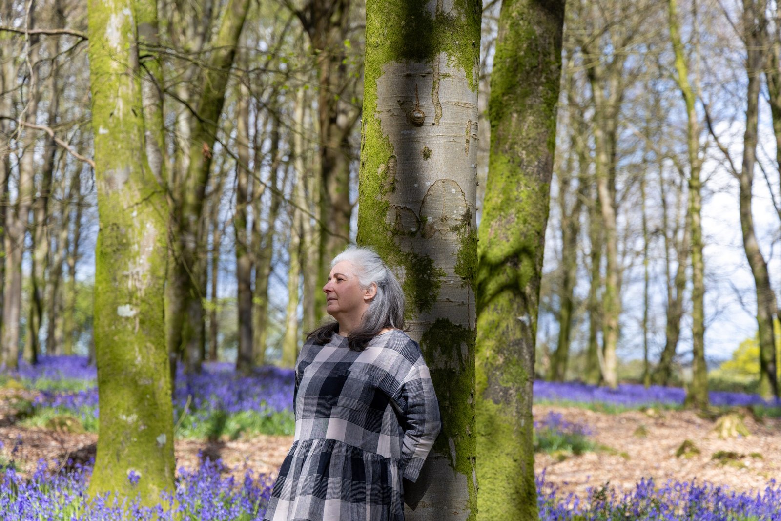 Arty Genny is in a forest, leaning against a tree trunk. You can see the bluebells in the background. Her facial expression is calm and content.