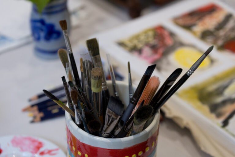 A brightly-painted ceramic pot sitting in the foreground on a table surface next to an open sketchbook showing paintings and there are also some pencils laid alongside. The ceramic pot is filled with a variety of paintbrushes