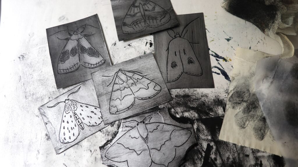 Inked Tetra Pak plates incised with various moth designs strewn on a messy, inky table