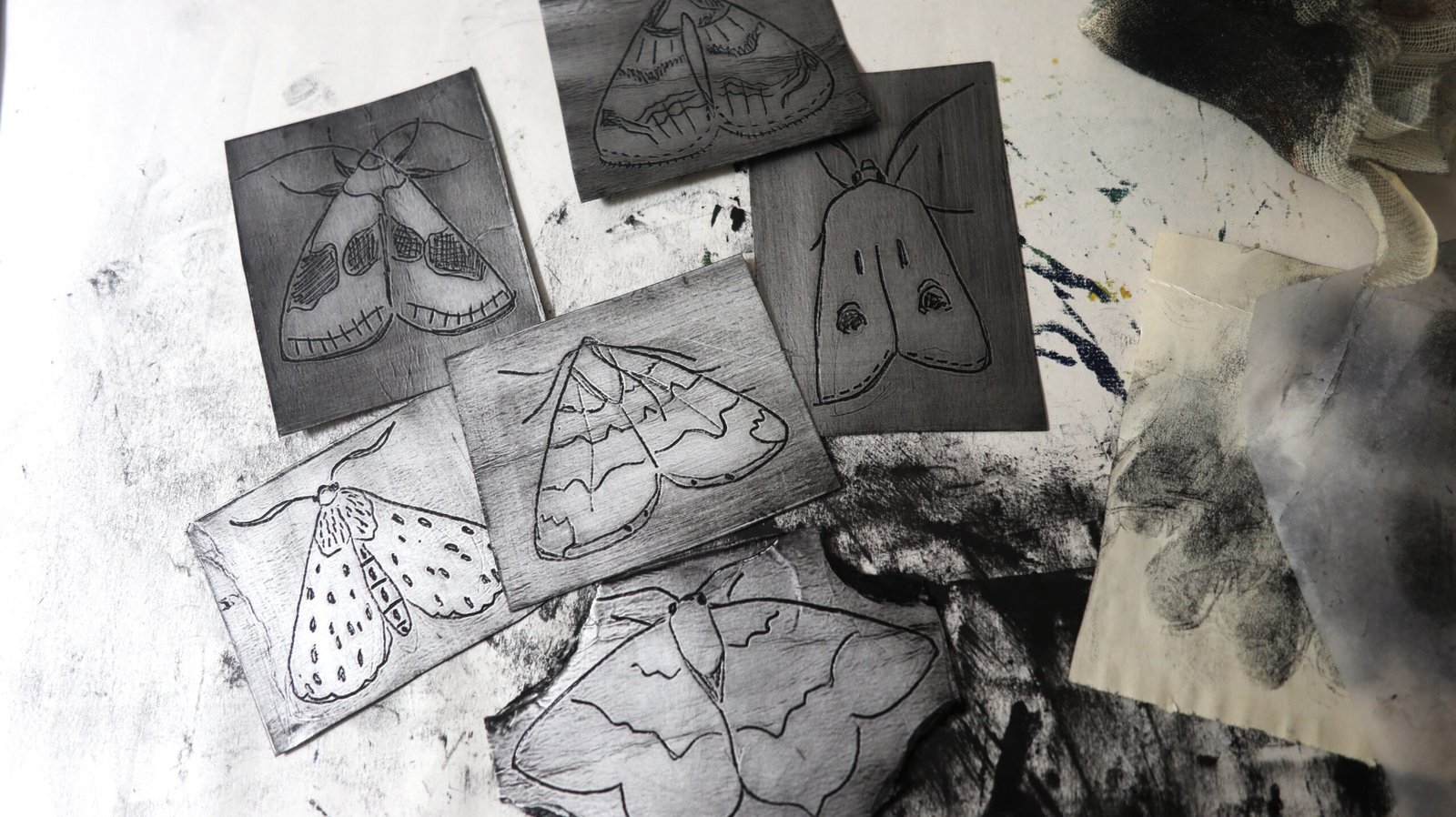 Inked Tetra Pak plates incised with various moth designs strewn on a messy, inky table