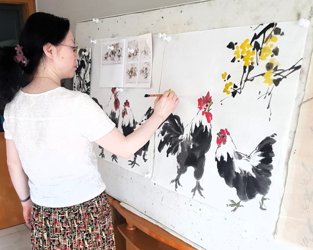 A female Chinese brush artist standing in front of a magnetic board, her right arm raised to paint. She is holding a Chinese brush and is adding detail to her Chinese ink painting of a rooster
